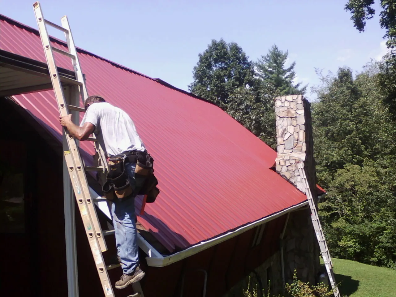 Roofer on a ladder beside a red metal roof and stone chimney — Soffit & Fascia Repair by Roofing Brothers in Laurelville